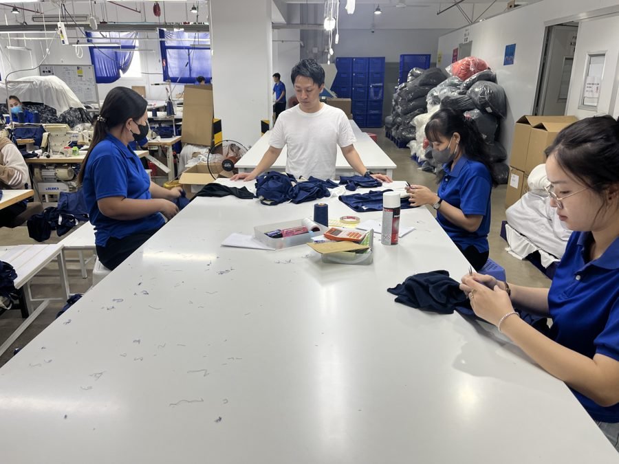 自社縫製工場の検品台で、スタッフが製品を 1 枚ずつ手に取って検品している様子
